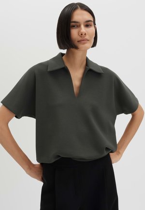 Vrouw met kort donker haar, die een losse donkergroene korte mouwen blouse met kraag draagt, staat met haar handen in de zij tegen een effen achtergrond.