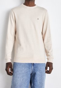 Beige långärmad tröja i mjukt stickat material, med rundad halsringning och ribbade ärmslut, prydd med en liten logotyp på bröstet. Bärs tillsammans med blå jeans.