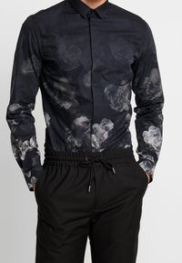 Musta pitkähiha teeppari, jossa on harmaan sävyinen kukkakuosi, napillinen kaulus ja slim fit -leikkaus. Yhdistetty mustiin joustaviin vyötäröhousuihin.