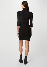 Robe noire côtelée avec des manches trois-quarts et un col montant. Présente des épaules puffées et une silhouette ajustée. Portée avec des bottes noires épaisses.