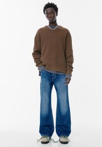 Brown geribbelde trui over een grijze lange mouwen shirt, gecombineerd met blauwe wijde jeans en beige sneakers met groene veters.