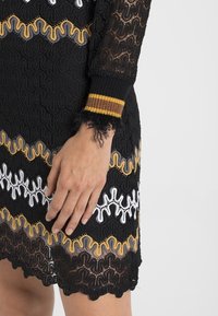 Svart spetsklänning med färgglada vågiga mönster i vitt och guld, med ribbade muddar i svart och gult samt en vågig fåll.