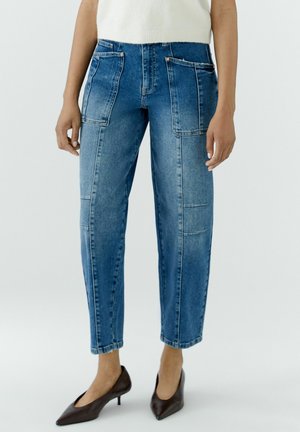 Vrouw die blauwe high-waist spijkerbroek draagt met grote opgestikte voorzakken en bruine kitten heels met puntige neus.