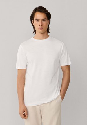 Joven con cabello oscuro de longitud media, vestido con una camiseta blanca de manga corta y pantalones beige claros, posando frente a un fondo neutro.