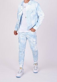 Světle modrá džínová bunda s tie-dye vzorem a stříbrnými knoflíky, doplněná o odpovídající cargo kalhoty. Pod ní je bílý tričko a šedé tenisky s modrými akcenty.