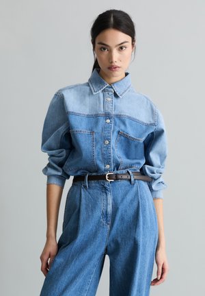 Femme aux cheveux foncés portant une chemise en denim bicolore bleu à boutons, rentrée dans un pantalon en denim taille haute à jambes larges, avec une ceinture noire.