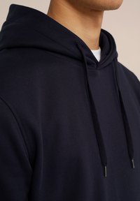 Tamsiai mėlynas džemperis su minkšta tekstūra, su raištelių gobtuvu ir metaliniais akcentais. Detalizuotas kadravimas, pabrėžiantis audinį ir dizaino ypatybes.