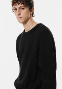 Maglione nero a maglia con texture a coste, scollatura ampia a girocollo e design con spalle cadenti, caratterizzato da una vestibilità larga e rilassata.
