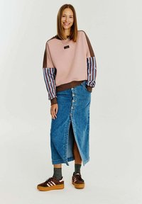 Hnedá a ružová mikina s pruhovanými rukávmi, skombinovaná s modrou džínsovou sukňou s predným zapínaním na gombíky a rozparkom. Hnedé tenisky.