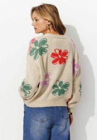 Pull en tricot beige avec des motifs floraux rouges, verts et roses. Présente une coupe décontractée, des manches à épaule tombante et un ourlet côtelé.