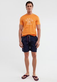 Oranžinė medvilninė marškinė su baltais "DENIM" grafikos elementais, derinama su tamsiai mėlynais maudymosi šortais su raudonu ir baltu raišteliu. Juodi šlepetės.