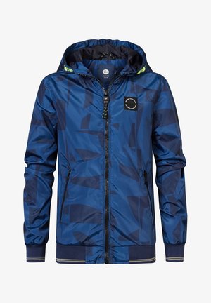 Enfant portant une veste zippée bleue à motifs, une chemise bleu clair, un jean retroussé et des baskets blanches, debout avec une main dans la poche.