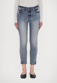 Világoskék, kopott hatású, skinny farmernadrág közepes derékkal, négy zsebbel és cropped szegéllyel, fekete lapos cipővel párosítva.