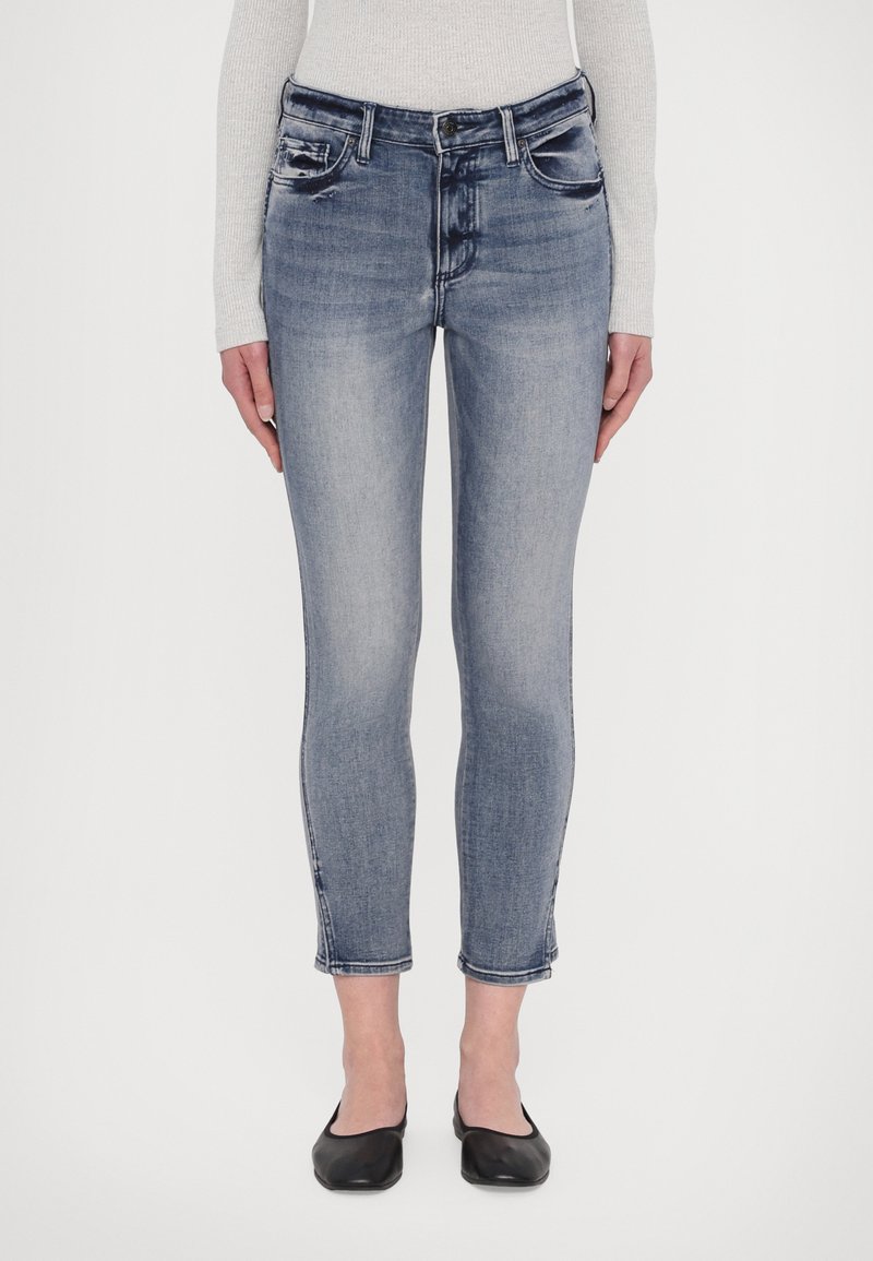 Világoskék, kopott hatású, skinny farmernadrág közepes derékkal, négy zsebbel és cropped szegéllyel, fekete lapos cipővel párosítva.
