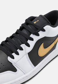 Sneaker in bianco e nero con tomaia in pelle texturizzata, presenta un logo dorato, lacci neri e suola in gomma. Visibile cucitura intricata.