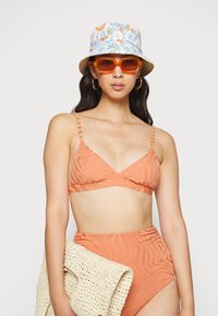 Biquíni laranja com padrão texturizado e ondulado; parte de baixo de cintura alta combinando. Acessórios incluem um chapéu tipo balde com flores e óculos de sol laranja.