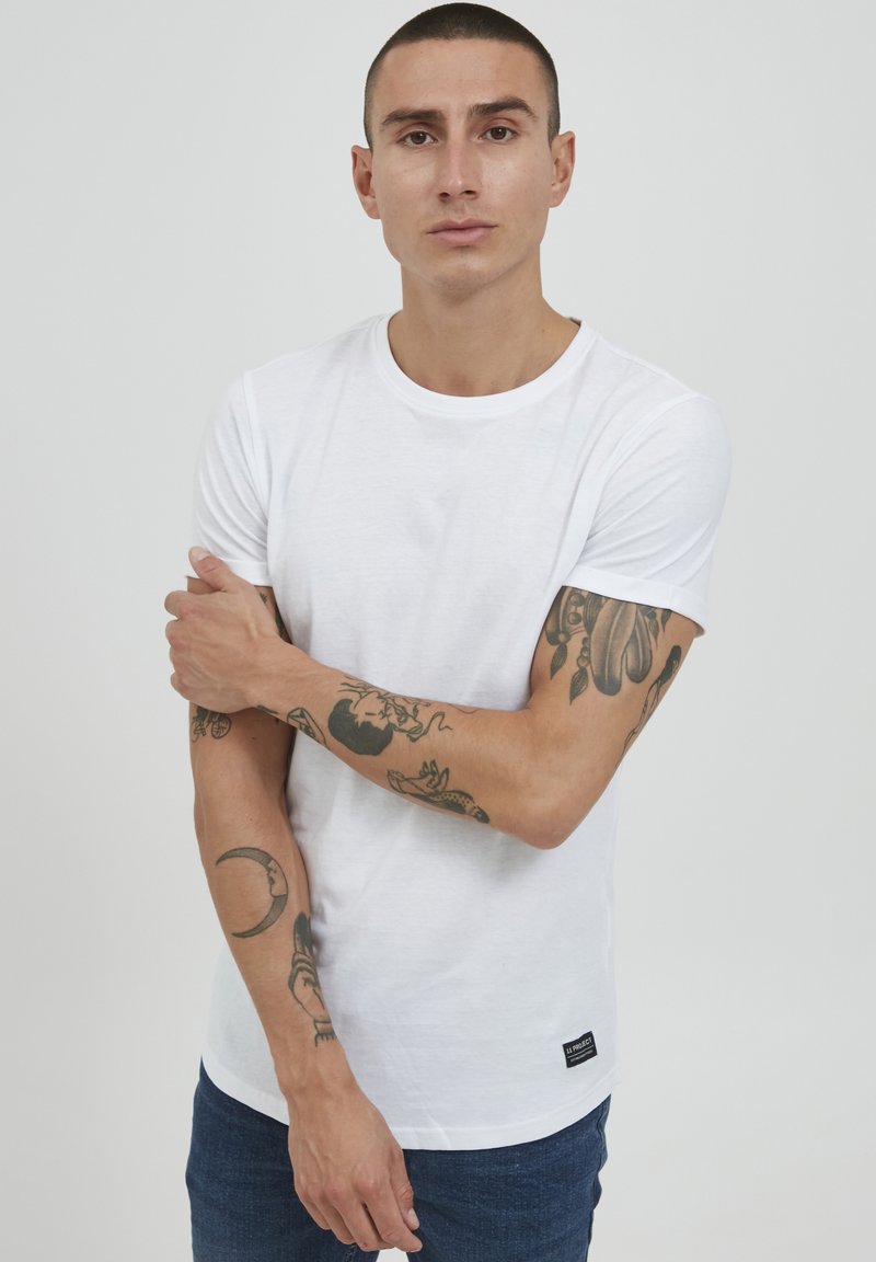 Hvid kortærmet t-shirt lavet af bomuld. Klassisk rund hals med en glat tekstur. Bæret med blå jeans og synlige tatoveringer på armene.