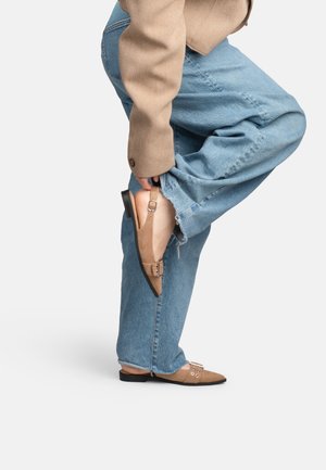 Personne portant un jean bleu clair et des ballerines beiges à bride arrière, levant une jambe pour ajuster la bride de la chaussure.