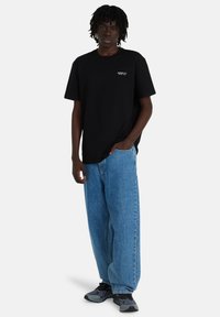 Young man standing wearing black t-shirt with "Element Co. Since 1992," baggy blue jeans, and black-gray sneakers against white background.