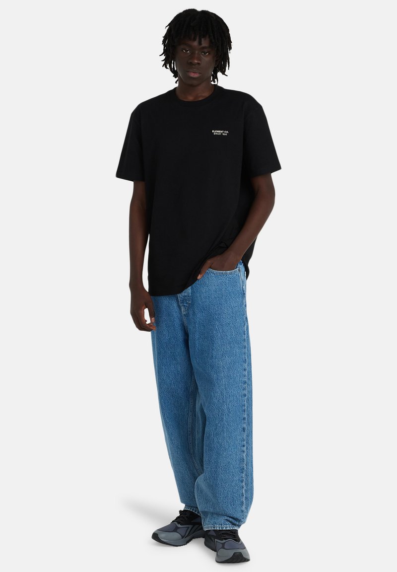 Young man standing wearing black t-shirt with "Element Co. Since 1992," baggy blue jeans, and black-gray sneakers against white background.