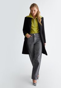 Cappotto nero, maglione verde brillante con collo zip e jeans wide-leg in tono argento. Il modello indossa tacchi metallici lucidi. Sfondo semplice.