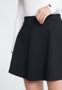 Zwarte flairrok met een gestructureerd ruitpatroon, hoge tailleband en zachte stof. Opgenomen van de taille naar beneden, vastgehouden met één hand.