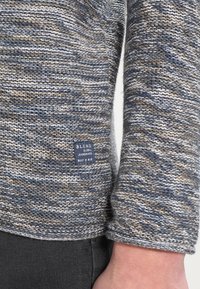 Pull en tricot dans des nuances de bleu, beige et gris avec des rayures texturées. Étiquette rectangulaire portant la mention "BLEND GLOBAL JEANMAKER."