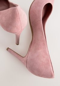 Een paar roze suède hoge hakken met spits toelopende neuzen en slanke hakken op een lichte ondergrond, getoond vanuit boven- en achterkant.