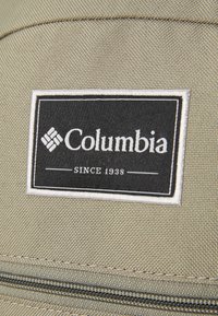 Columbia brand patch in black and white on beige fabric, featuring "SINCE 1938" text and a diamond logo, with textured stitching detail.