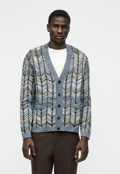 Άνδρας που φοράει μπλε-γκρι ζακέτα με κουμπιά και πολύχρωμα ζιγκ-ζαγκ μοτίβα πάνω από λευκό πουκάμισο και σκούρο καφέ παντελόνι.