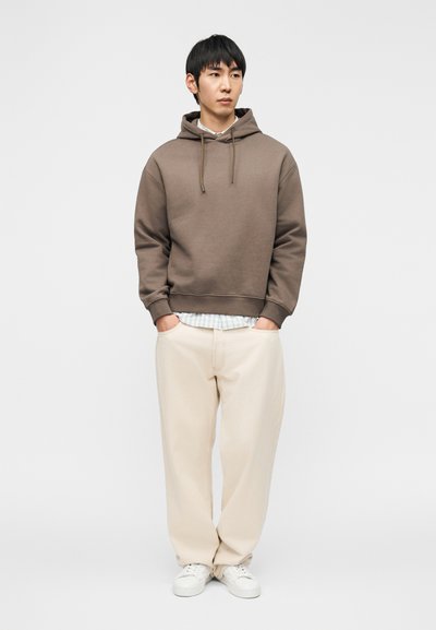 Νέος άνδρας όρθιος με τα χέρια στις τσέπες, φοράει καφέ φούτερ με κουκούλα, ανοιχτόχρωμο πουκάμισο με καρό σχέδιο, κρεμ φαρδιά παντελόνια και λευκά αθλητικά παπούτσια σε λευκό φόντο.