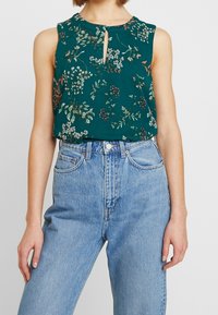 Haut sans manches à motifs floraux verts avec un détail en forme de trou à l'avant, confectionné dans un tissu léger, associé à un jean en denim taille haute.