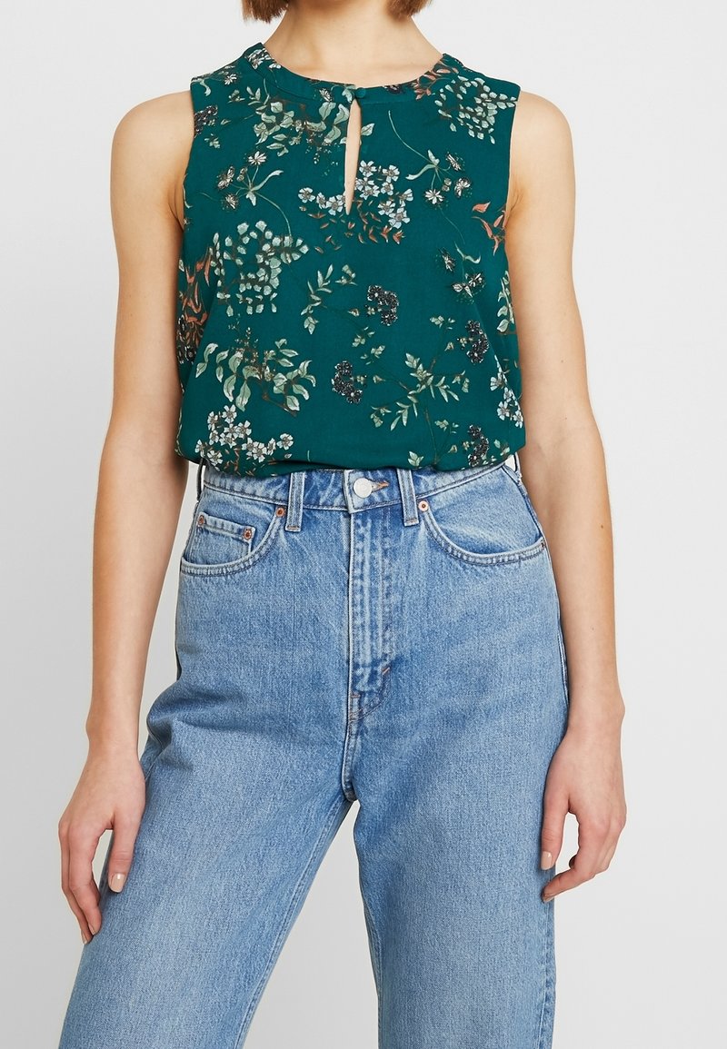 Haut sans manches à motifs floraux verts avec un détail en forme de trou à l'avant, confectionné dans un tissu léger, associé à un jean en denim taille haute.