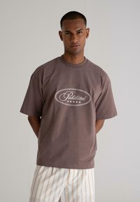 Brown oversized t-shirt in soft cotton with a round neck and short sleeves, featuring a white graphic print that reads "Prohibited."