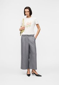 Femme portant un t-shirt blanc à fleurs "PEONIES", un pantalon large gris cropped, des talons slingback noirs pointus, et portant une sangle de sac à dos vert clair.