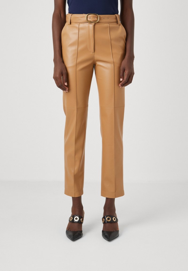 Mannequin portant un pantalon en cuir camel taille haute avec une ceinture assortie et des escarpins noirs à bout pointu avec des détails d'œillets dorés.