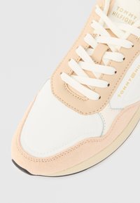 Λήψη από κοντά ενός μπεζ, λευκού και ροδακινί αθλητικού παπουτσιού Tommy Hilfiger με λευκές κορδόνες και το όνομα της μάρκας ορατό στη γλώσσα και στη πλευρά.