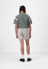 Chemise verte à manches courtes avec des rayures bleues et jaunes, associée à un short gris. Des chaussures noires et des chaussettes blanches complètent la tenue.
