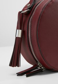 Rund, burgundy håndtaske lavet af tekstureret materiale; har en sølv lynlås, sort trim og en dekorativ kvast.