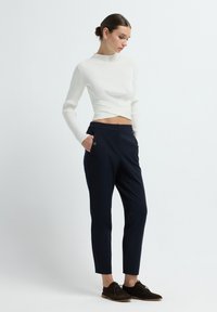 Vrouw in een witte geribbelde lange mouw crop top, marineblauwe op maat gemaakte broek met knoopzakken en zwarte veterschoenen, staand met haar handen in de zakken.