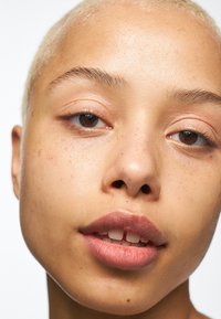 Gros plan sur le visage d'une personne avec des cheveux blonds clairs et courts. Ses traits incluent une peau lisse, des joues couvertes de taches de rousseur et une couleur de lèvres naturelle avec une légère brillance.
