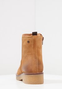 Stivale alla caviglia in suede tan con punta rotonda, chiusura con zip e suola in gomma testurizzata. Cuciture marroni e un piccolo dettaglio del logo sul retro.