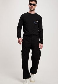 Czarna koszula z długim rękawem z kieszonką, zestawiona z czarnymi spodniami cargo i białymi sneakersami. Koszula ma teksturowany wzór i metkę marki.