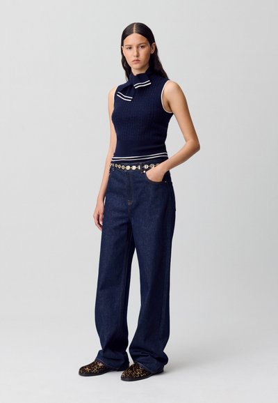 Mujer vestida con un top de punto sin mangas en color azul marino con cuello, jeans de mezclilla oscuros de cintura alta, un cinturón de monedas y zapatos de estampado de leopardo, de pie con la mano en el bolsillo.