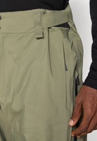 Pantaloni de culoare verde măslin cu o textură netedă, având un talie cu curea, buzunare cu fermoar și detalii de cusătură rezistente.