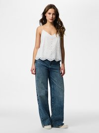 Mladá žena stojí, má na sobě bílý vyšívaný vršek bez rukávů, široké modré džíny a bílé tenisky, na jednobarevném světlém pozadí.