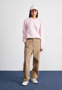 Felpa rosa chiaro con logo bianco, pantaloni cargo beige con tasche laterali e scarpe da ginnastica bianche con dettagli rossi. Il cappellino è beige chiaro.