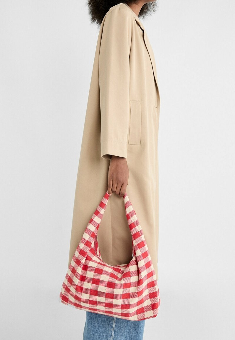 Personne portant un manteau beige et un jean bleu tenant un sac à main en tissu à carreaux rouges et blancs par sa poignée à son côté.