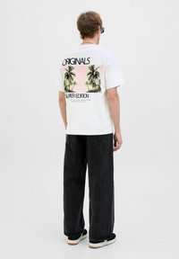 Hvid bomulds t-shirt med palmetræ grafik og teksten "ORIGINALS SILVER EDITION" på ryggen, parret med sorte brede bukser og sneakers.