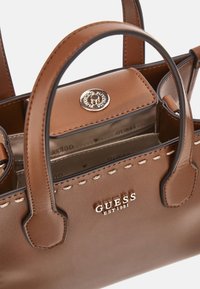 Borsa a mano in pelle marrone con due manici superiori, dotata di hardware dorato, logo GUESS e dettagli di cucitura sui lati. L'interno presenta più scomparti.